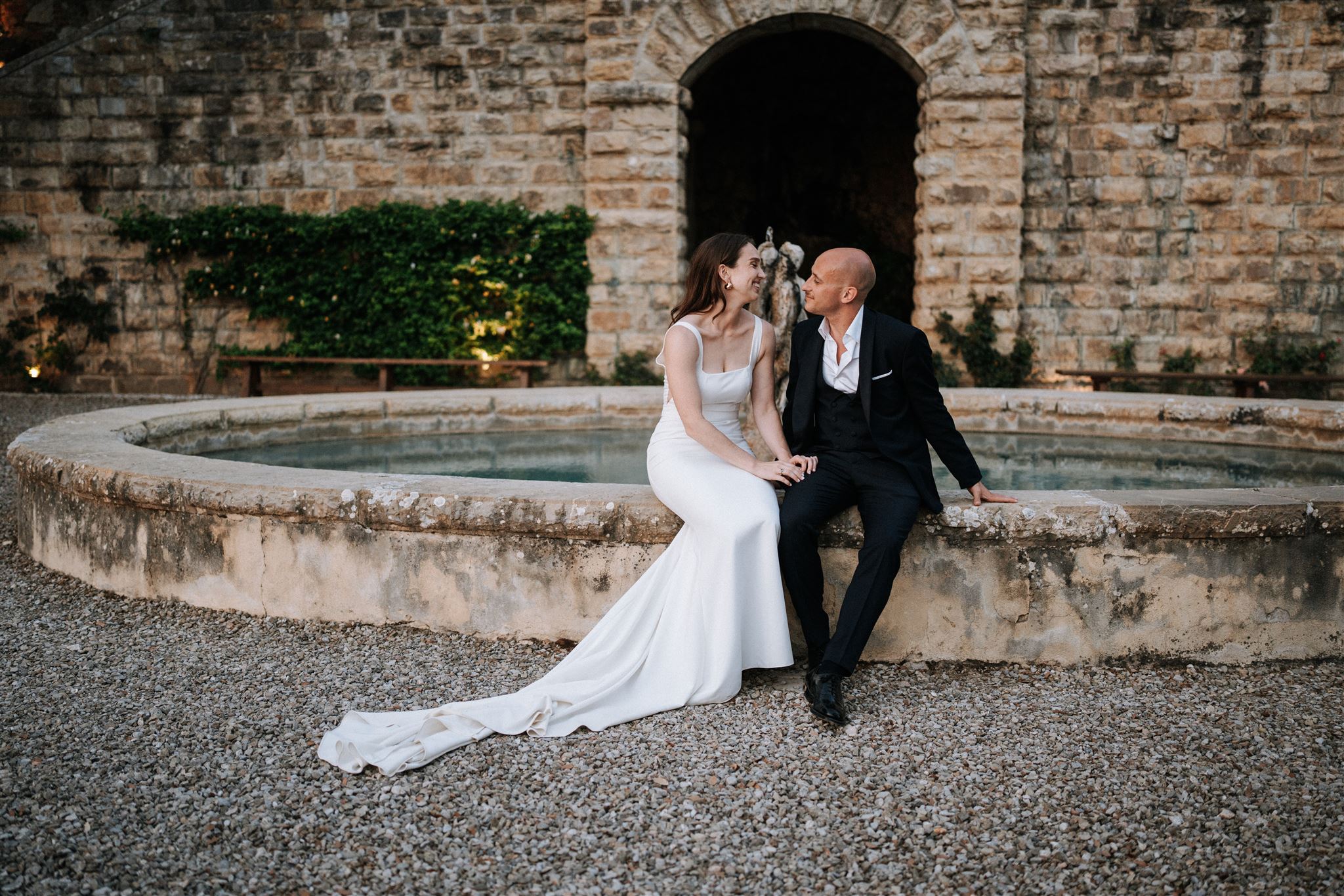 Bride and groom sitting together on an ancient stone fountain at their Tuscany wedding, photographed by Meredith Lord, destination wedding photographer