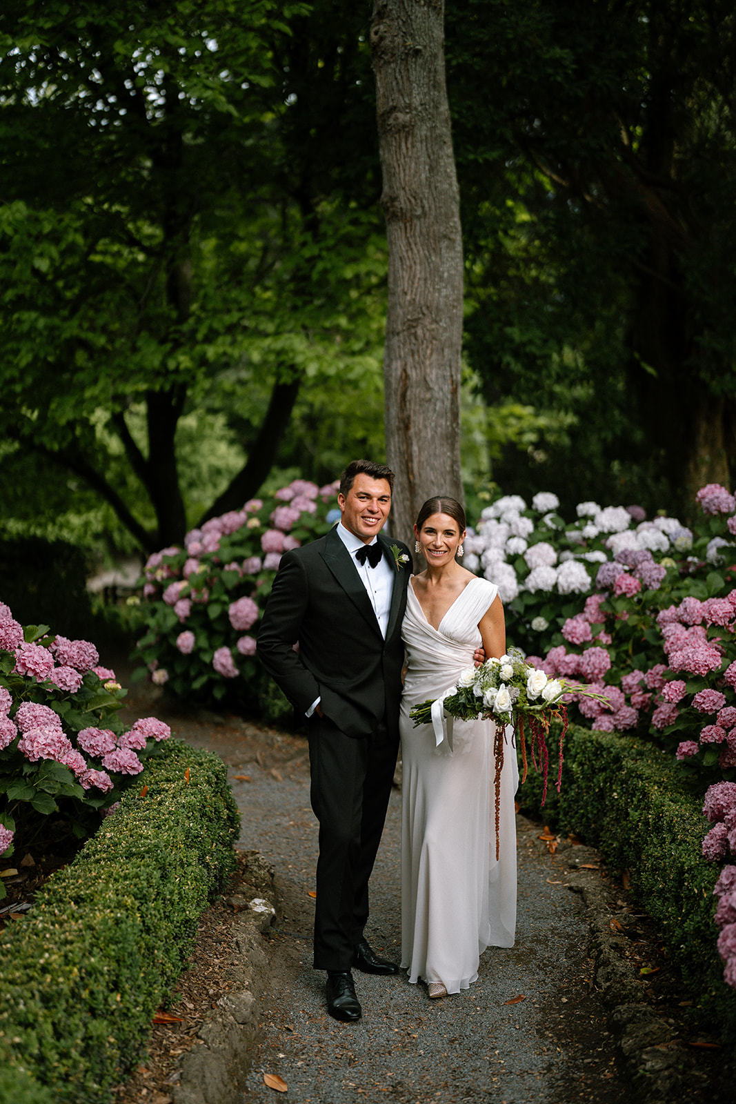 Georgie & James, Garden Wedding, New Zealand - Meredith Lord Photography Bride and groom pose together on a garden path framed by pink and white hydrangeas.