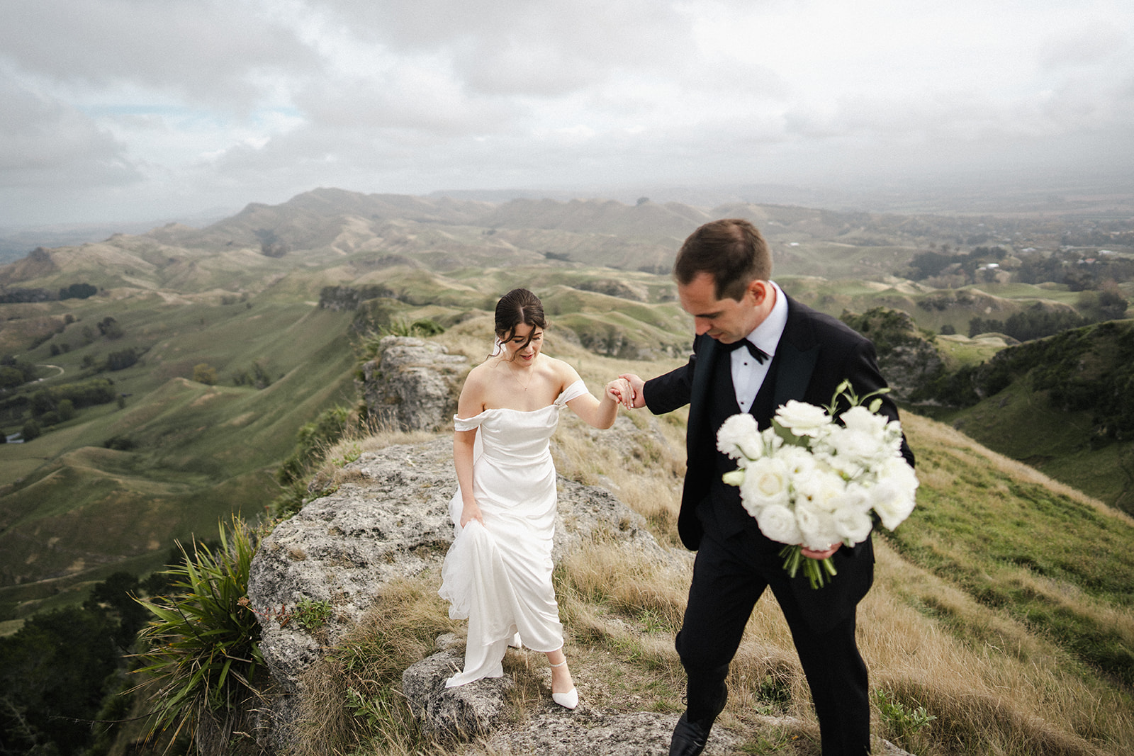 Maggie & Christo - Black Barn Wedding, Hawke's Bay - Meredith Lord Photography Groom leading his bride over a rocky mountain ridge
